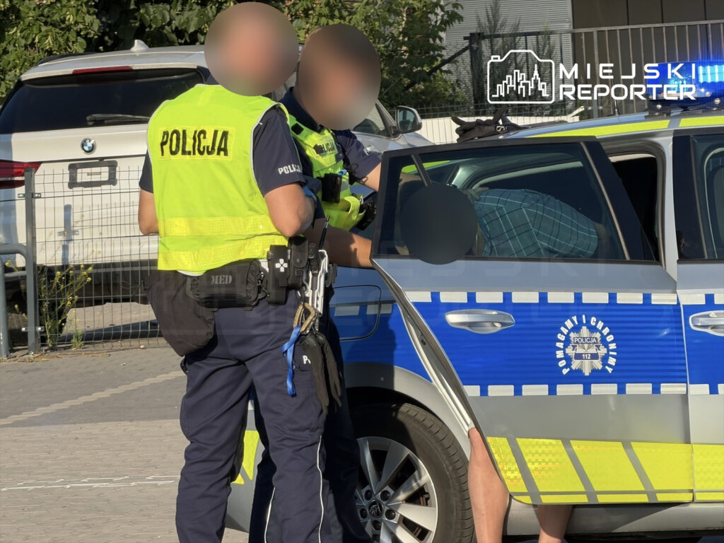 Policjanci w odblaskowych kamizelkach rozmawiają z mężczyzną siedzącym w radiowozie na parkingu.