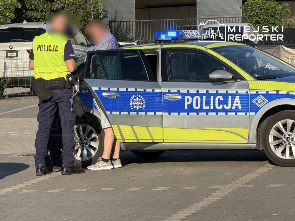 Policjant w kamizelce odblaskowej rozmawia z mężczyzną stojącym obok radiowozu policyjnego na parkingu.