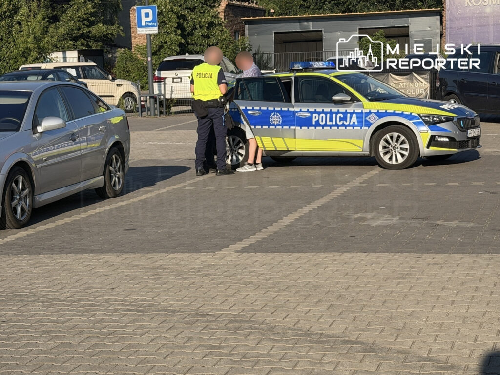 Policjant w kamizelce odblaskowej rozmawia z mężczyzną w krótkich spodenkach obok radiowozu na parkingu.