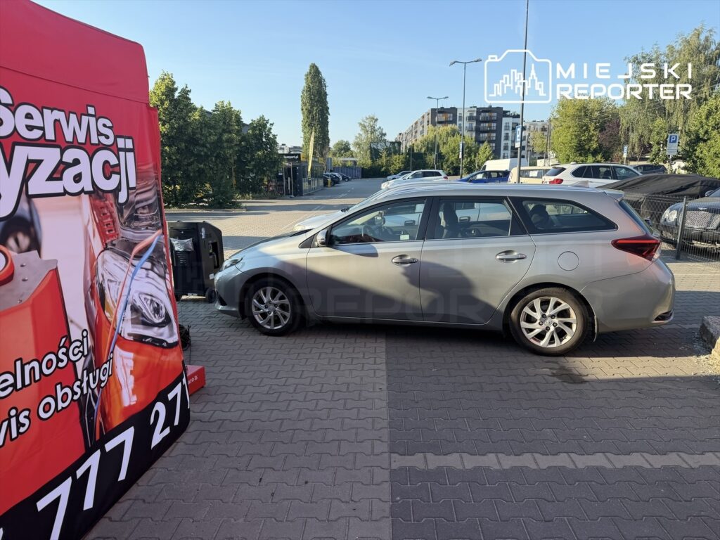 Szary samochód osobowy zaparkowany obok stoiska z usługami serwisu samochodowego w słoneczny dzień.