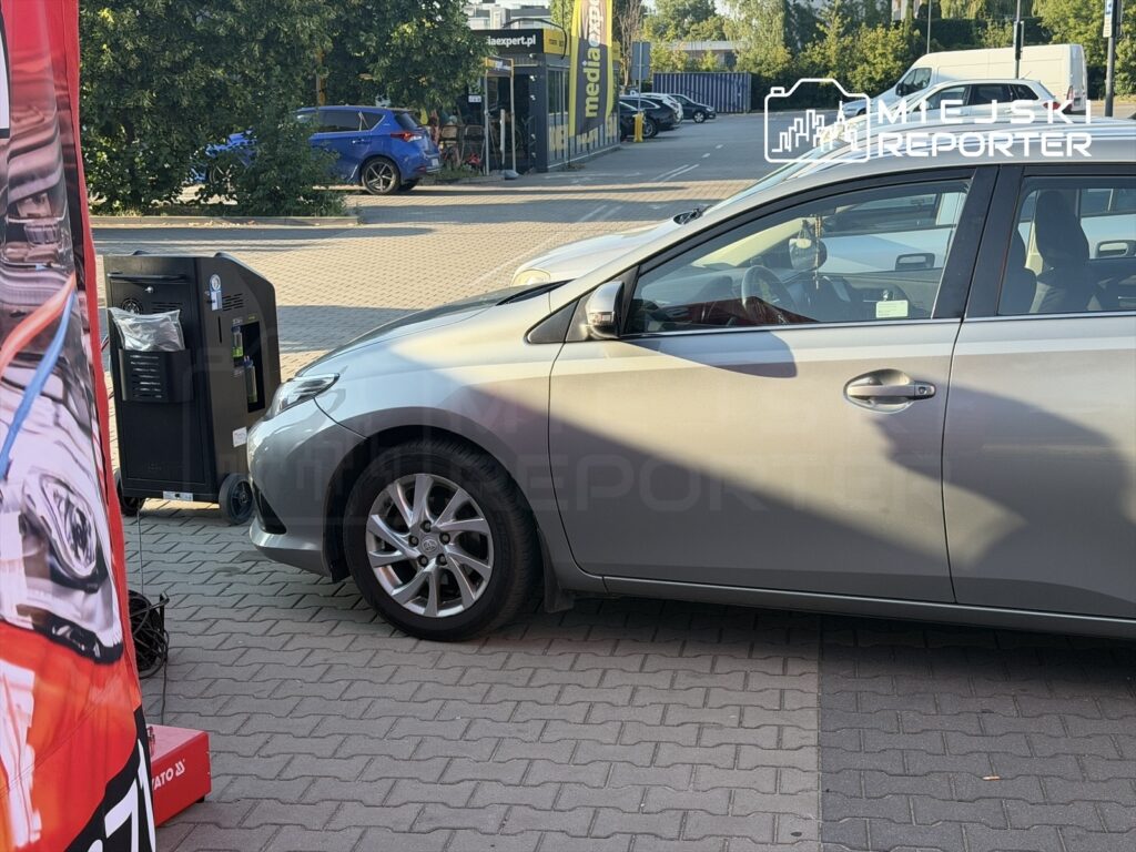 Samochód osobowy zaparkowany na stacji obsługi, obok urządzenia do nabijania klimatyzacji. W tle widać inne pojazdy.