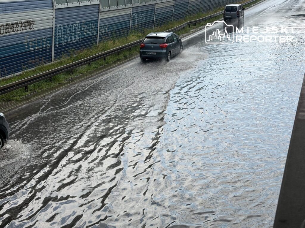 Samochody poruszają się po zalanej drodze, tworząc fale na wodzie, w otoczeniu metalowych ogrodzeń.