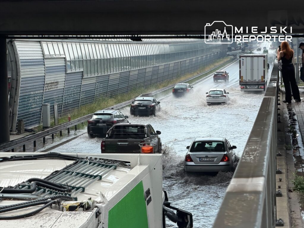 Samochody poruszają się po zalanej trasie S8, obok nowoczesnej przeszklonej ściany. Na poboczu stoi kobieta.