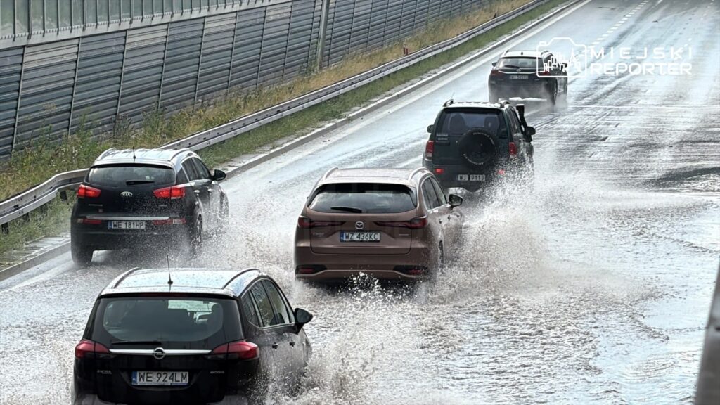 Samochody poruszają się po zalanej drodze, rozpryskując wodę na trasie S8. W tle widoczna jest wysoka bariera dźwiękochłonna.