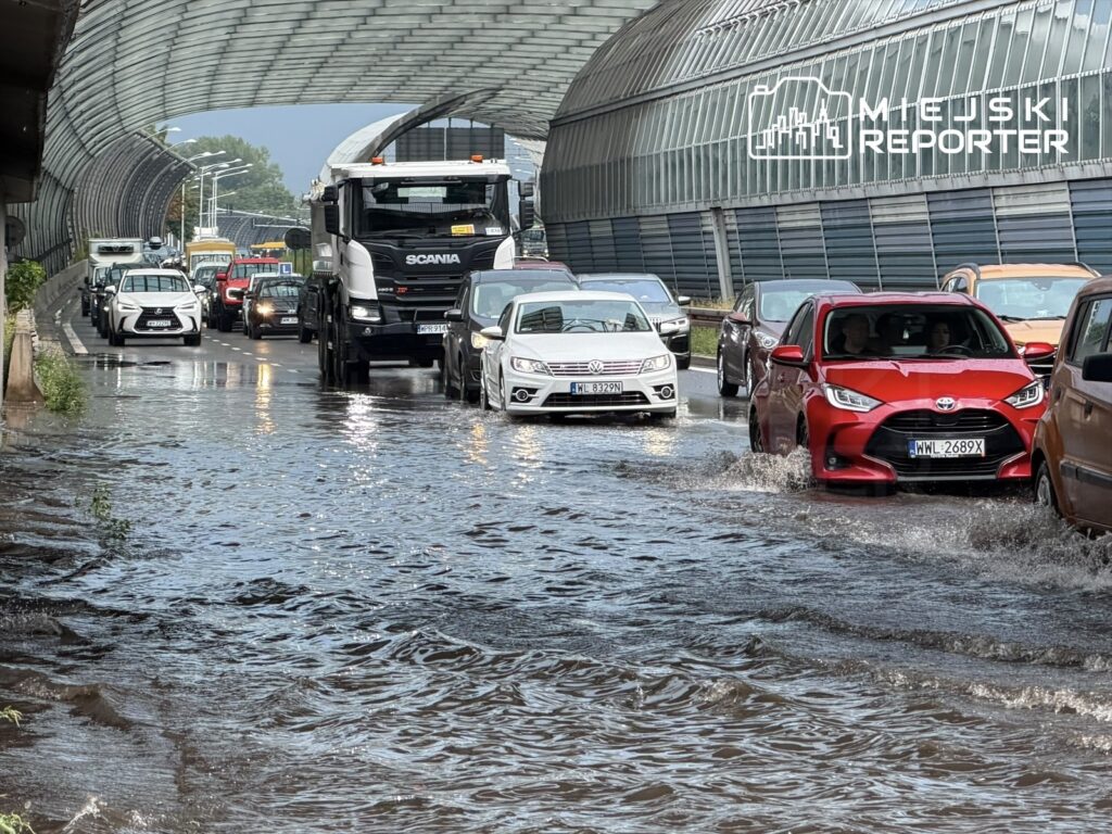 Ruch samochodowy na zalanej ulicy, z wodą sięgającą do kół pojazdów, w otoczeniu nowoczesnych budynków.