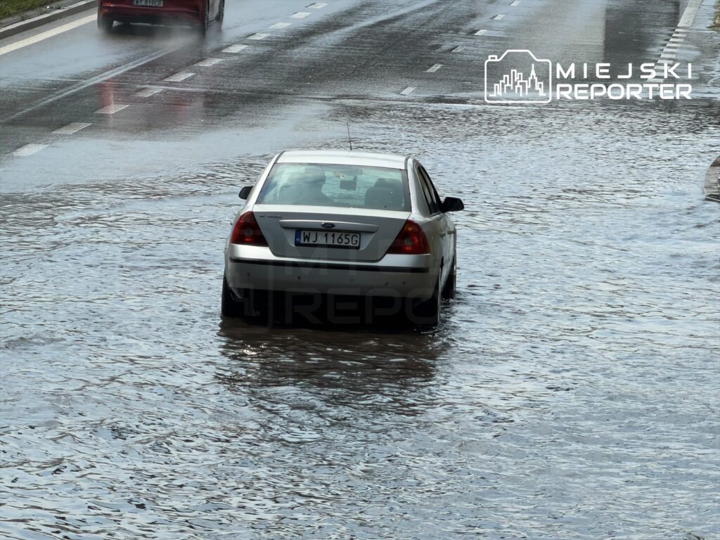 Samochód osobowy porusza się po zalanej drodze, woda odbija światło słoneczne, tworząc mokrą nawierzchnię.
