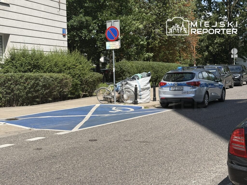 Policyjny radiowóz zaparkowany na niebieskim miejscu postojowym, obok rower i worki z odpadami, otoczenie zieleni.