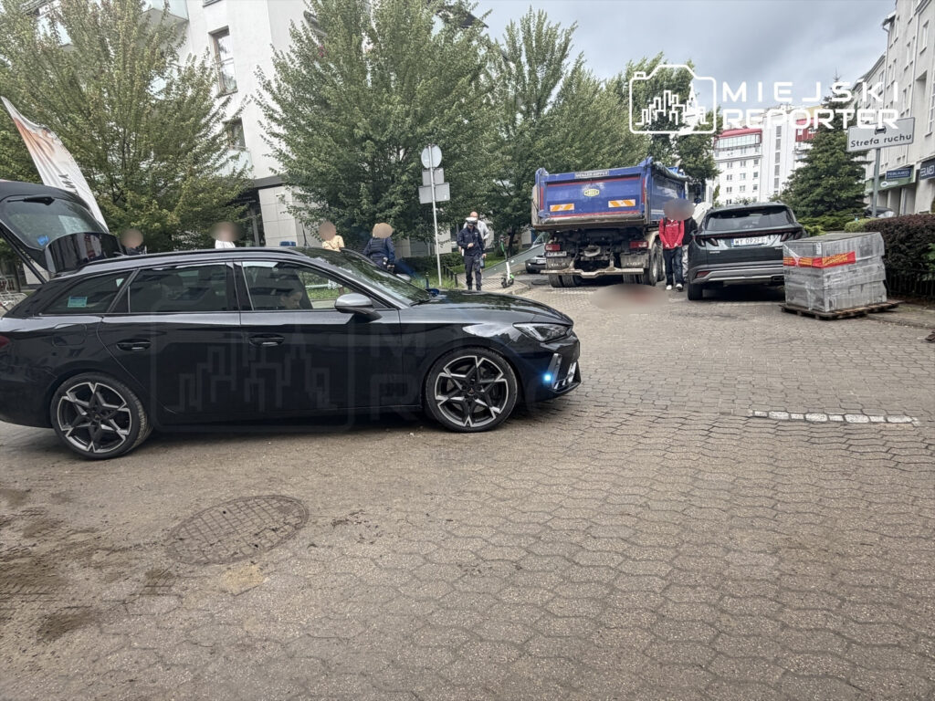 Czarny samochód osobowy stoi na drodze obok ciężarówki, a w tle widać grupę ludzi oraz budynki otoczone zielenią.