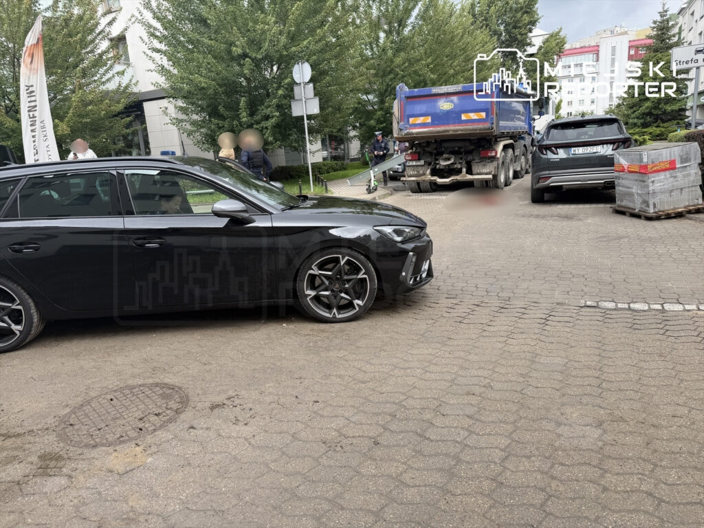 Czarny samochód osobowy zaparkowany na chodniku obok ciężarówki, w tle widoczni przechodnie oraz zieleń drzew.