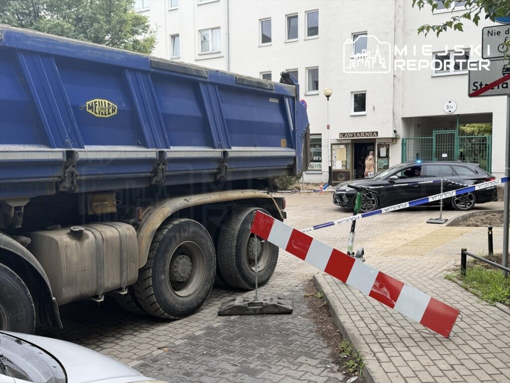 Ciężarówka z niebieską skrzynią stoi na ulicy, obok oznaczenia policyjnego i blokady drogi, w tle kawiarnia.