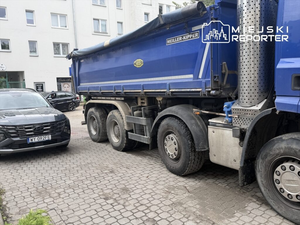Ciężarówka z niebieską naczepą stoi na wąskiej ulicy obok zaparkowanego czarnego samochodu osobowego.
