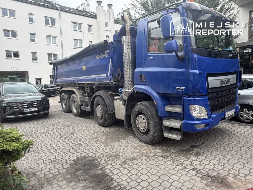 Niebieski ciężarowy pojazd marki DAF zaparkowany na kostkowanej nawierzchni obok ciemnego samochodu osobowego. W tle widoczny biały budynek mieszkalny.