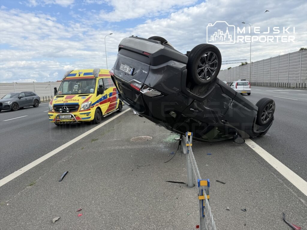 Na autostradzie stoi przewrócone na dach czarne auto, obok zatrzymana karetka pogotowia.