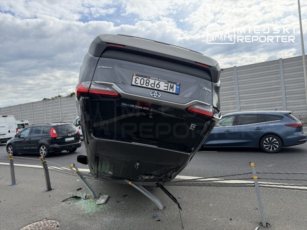 Na drodze leży przewrócony samochód osobowy na boku, otoczony przez inne pojazdy.