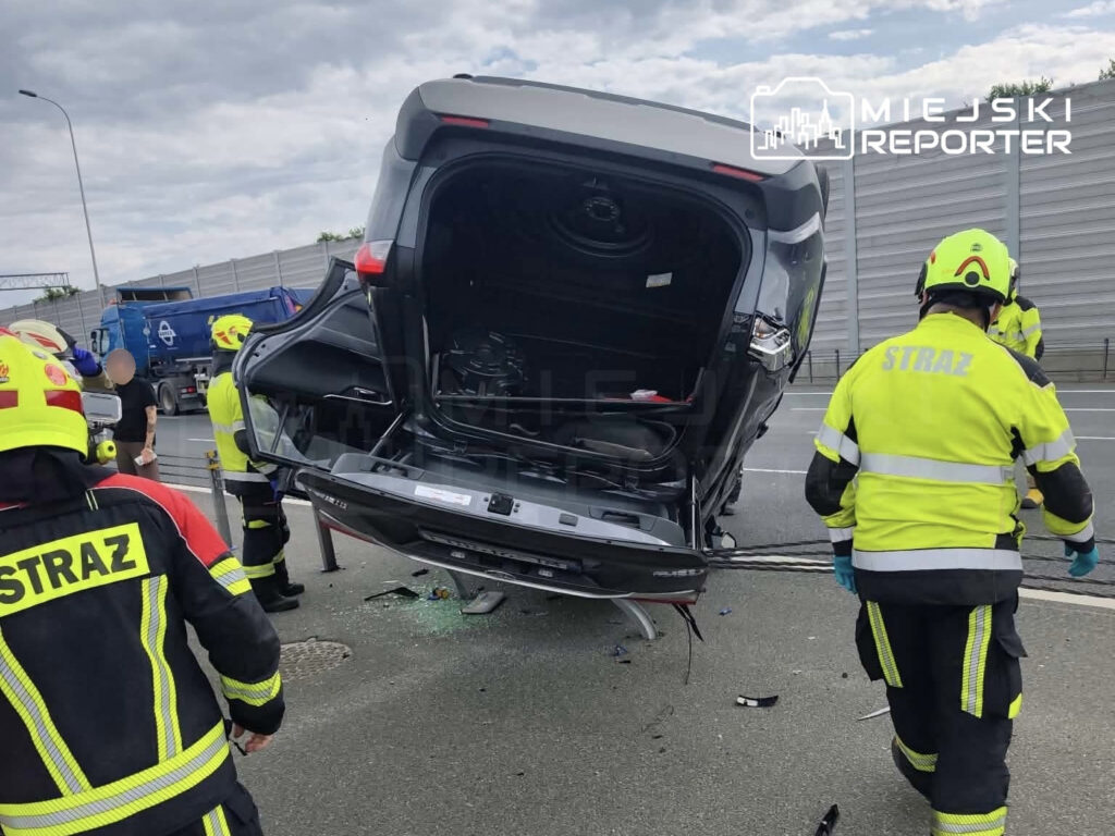 Toyota leży na dachu przy zjeździe z węzła, a strażacy w żółtych kaskach udzielają pomocy.