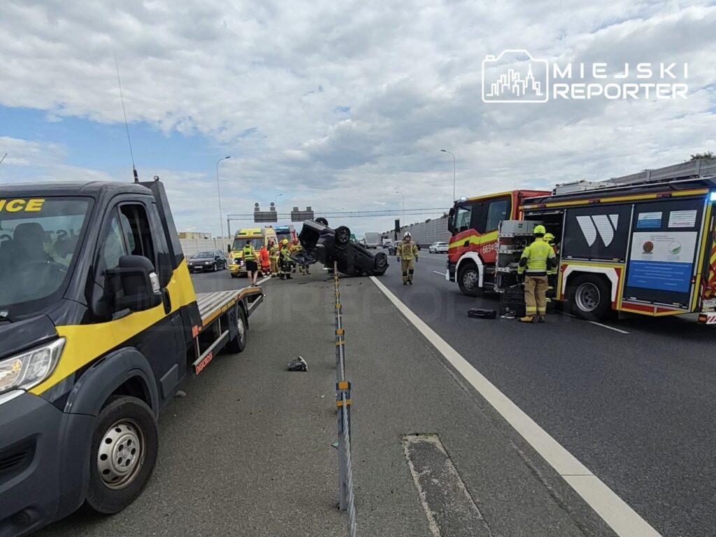 Na zjeździe z węzła drogi stoi przewrócony samochód, obok pracują strażacy i ratownicy medyczni. W tle widać pojazdy służb ratunkowych.