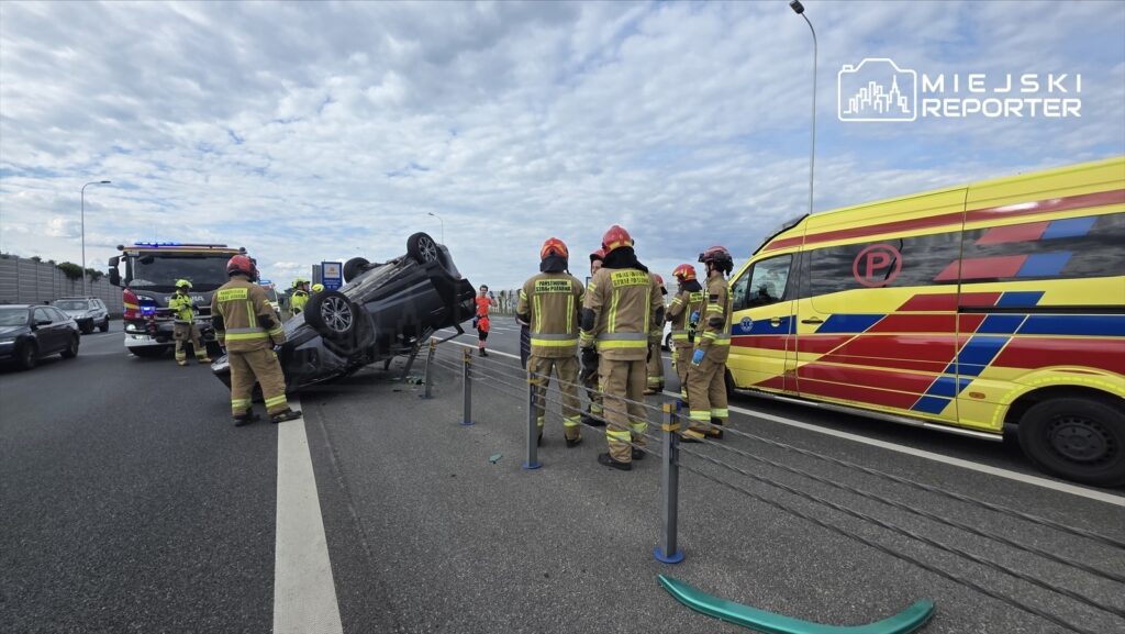 Grupa strażaków w mundurach i kaskach otacza przewrócony samochód osobowy na pasie ruchu. W tle widać wóz strażacki oraz ambulans.