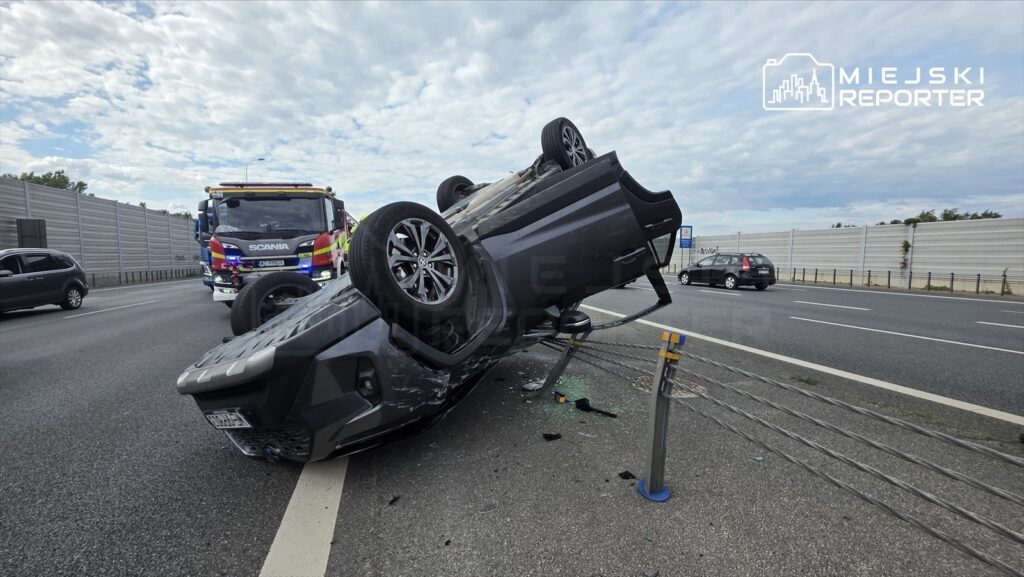 Czarny SUV leży na dachu przy drodze, obok widoczny wóz strażacki oraz inne pojazdy w tle.