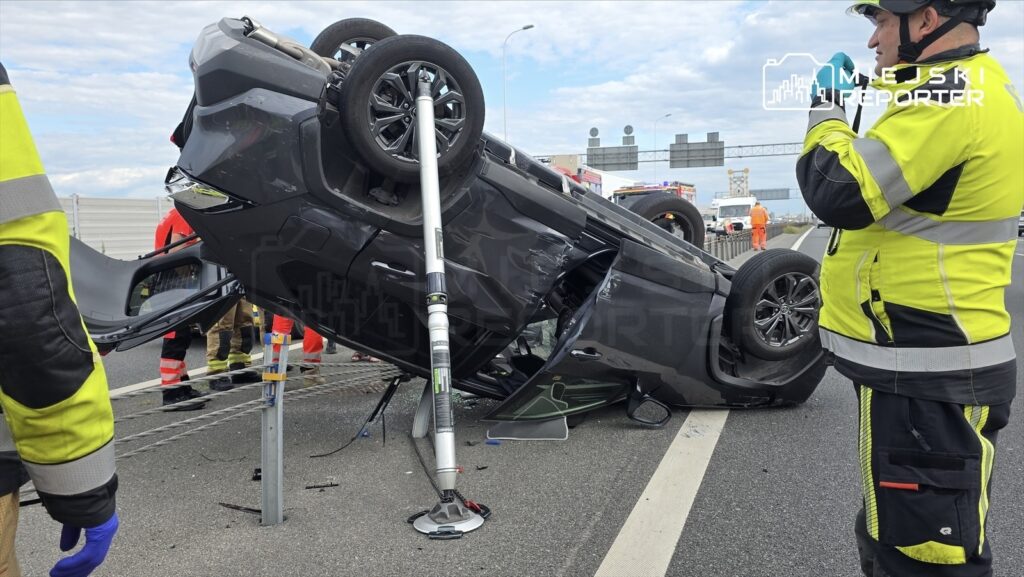 Toyota dachująca na jezdni, wokół niej strażacy w żółtych mundurach i służby ratunkowe.