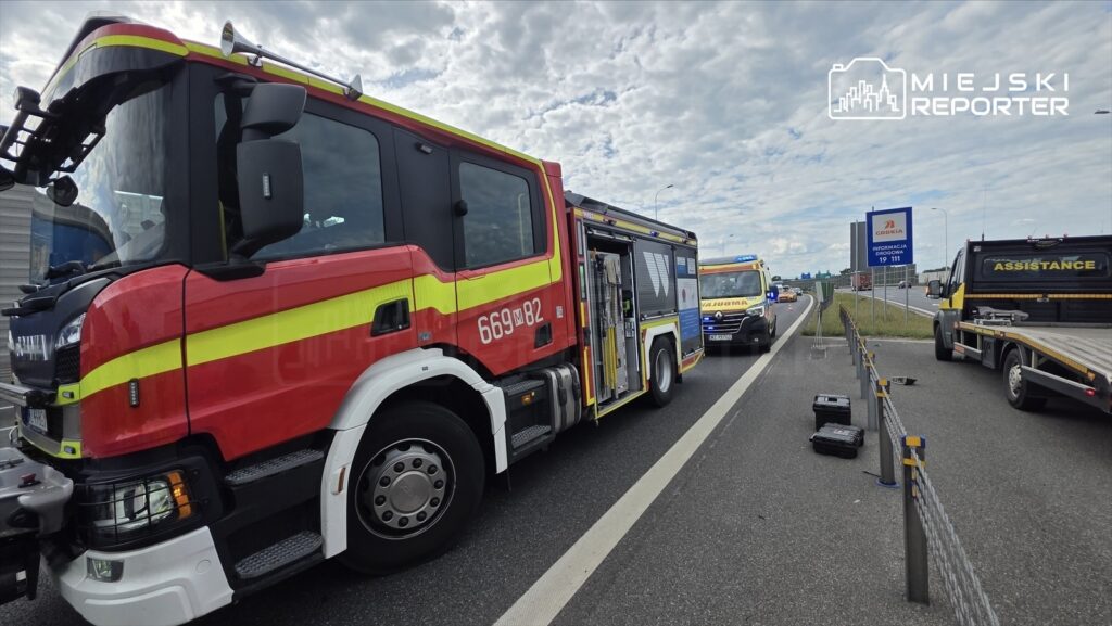 Czerwony wóz strażacki z widoczną numeracją 669M82 stoi na poboczu drogi, obok niego niebieski samochód z sygnalizacją.