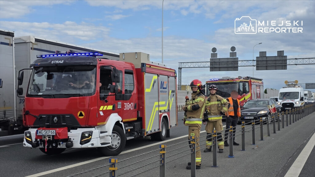 Wóz strażacki zatrzymany na autostradzie, obok strażacy w mundurach rozmawiają z kierowcą.