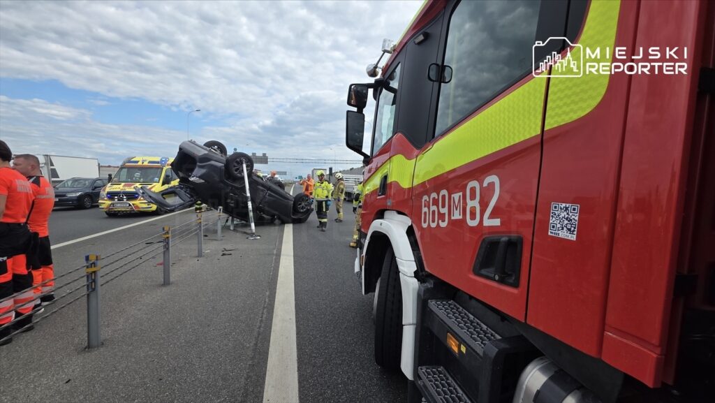 Na jezdni leży przewrócone auto, w tle zespół ratowników i straż pożarna w akcji.