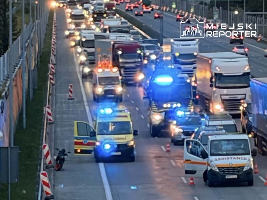 Karetka pogotowia z włączonymi sygnałami świetlnymi zatrzymana przy zablokowanej drodze, obok leżący motocykl.