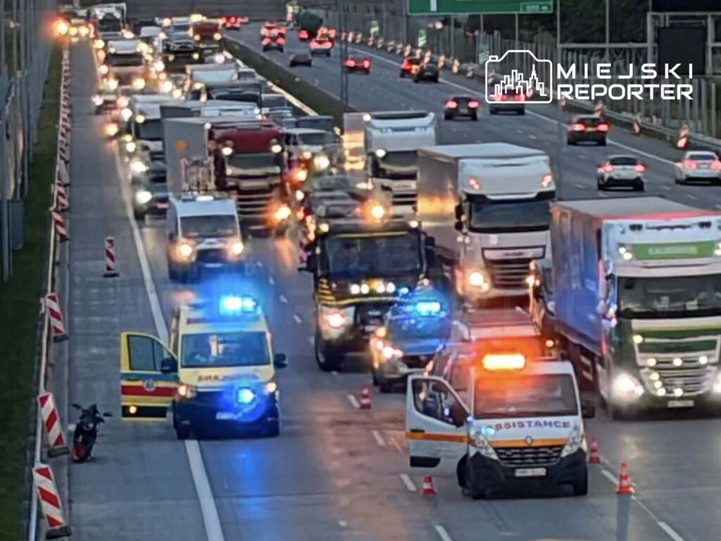 Karetka z włączonymi sygnałami świetlnymi zatrzymana na drodze, obok niej widać zablokowany ruch pojazdów ciężarowych.