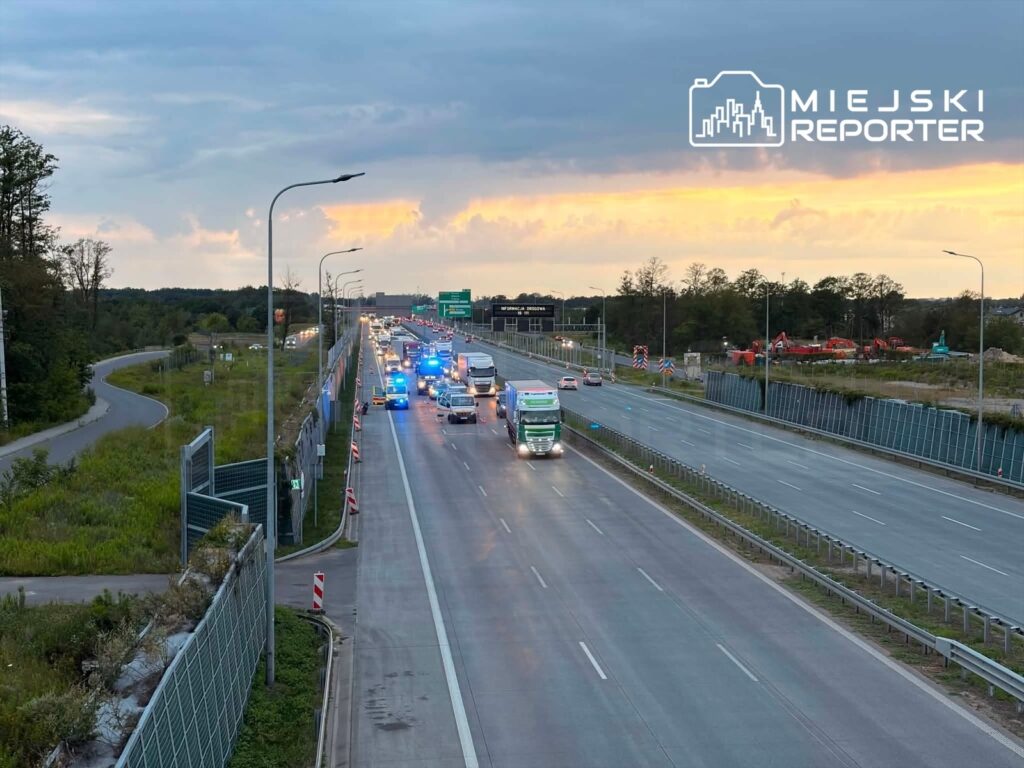 Zespół pojazdów służb ratunkowych zatrzymany na drodze ekspresowej, w tle wschodzące słońce i pojazdy osobowe.