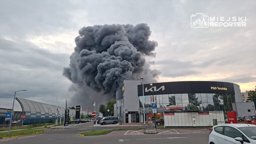 Gęste, czarne kłęby dymu unoszą się nad nowoczesnym salonem samochodowym, w tle widoczne szare niebo.