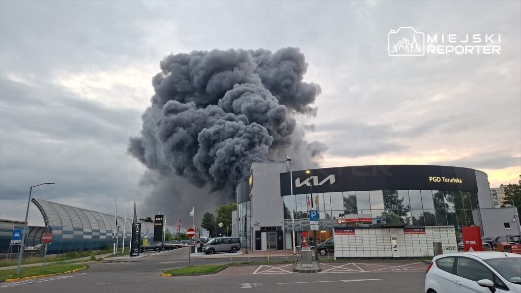 Kłęby czarnego dymu unoszą się nad nowoczesnym salonem samochodowym w mieście, w tle szare niebo.