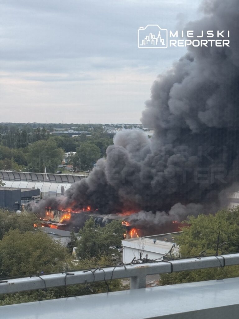 Ogromne kłęby czarnego dymu unoszą się nad płonącym salonem samochodowym otoczonym drzewami.