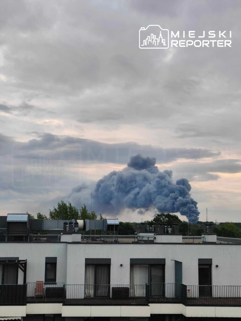 Kłęby ciemnego dymu unoszą się nad miastem, widoczne w tle za nowoczesnym budynkiem mieszkalnym.