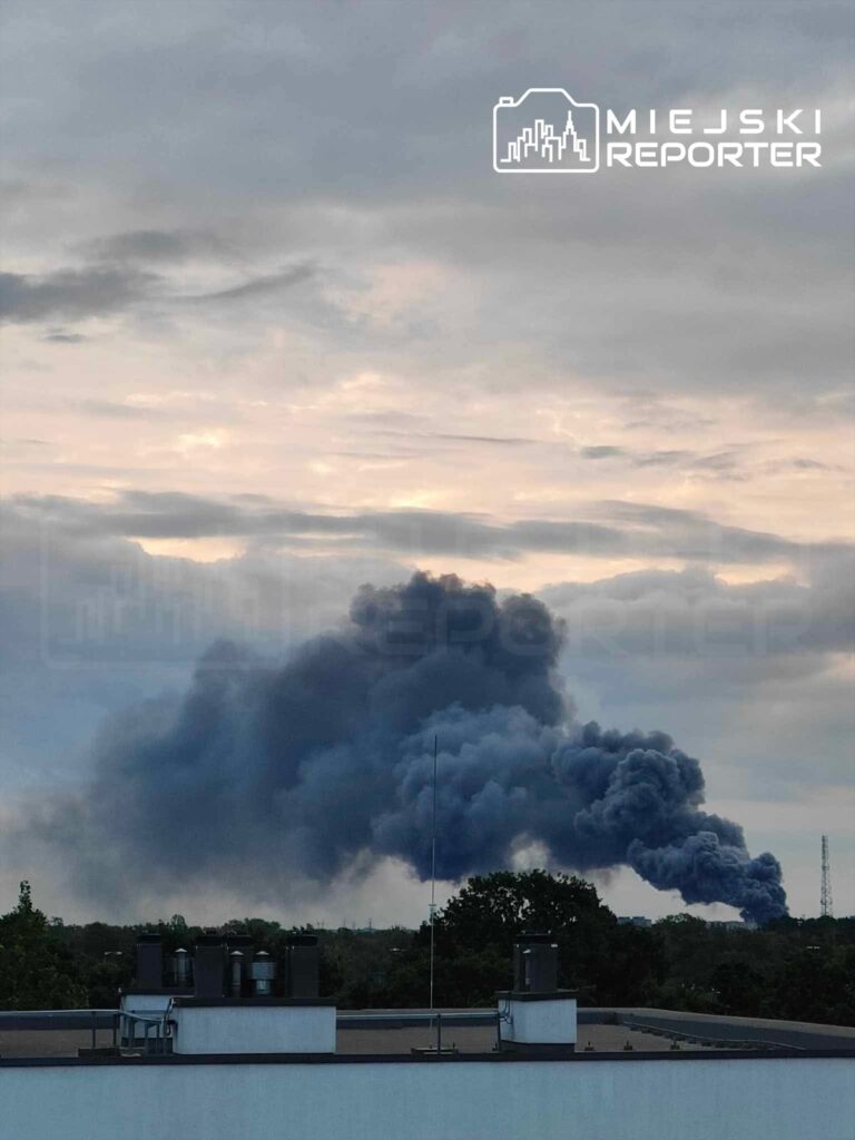 Kłęby czarnego dymu unoszą się nad horyzontem, widoczne za dachem budynku w miejskim otoczeniu.