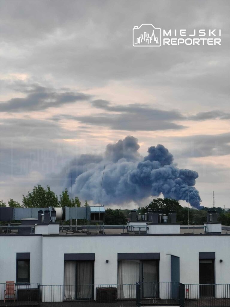 Gęste kłęby dymu unoszą się nad miastem, widoczne w tle za nowoczesnym budynkiem mieszkalnym.