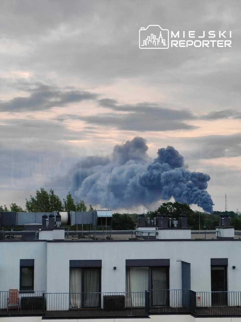 Kłęby czarnego dymu unoszą się nad miastem, widoczne w tle za nowoczesnym budynkiem mieszkalnym.