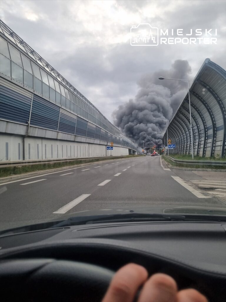 Kłęby czarnego dymu unoszą się nad miastem, widoczne z drogi prowadzącej obok nowoczesnego biurowca.