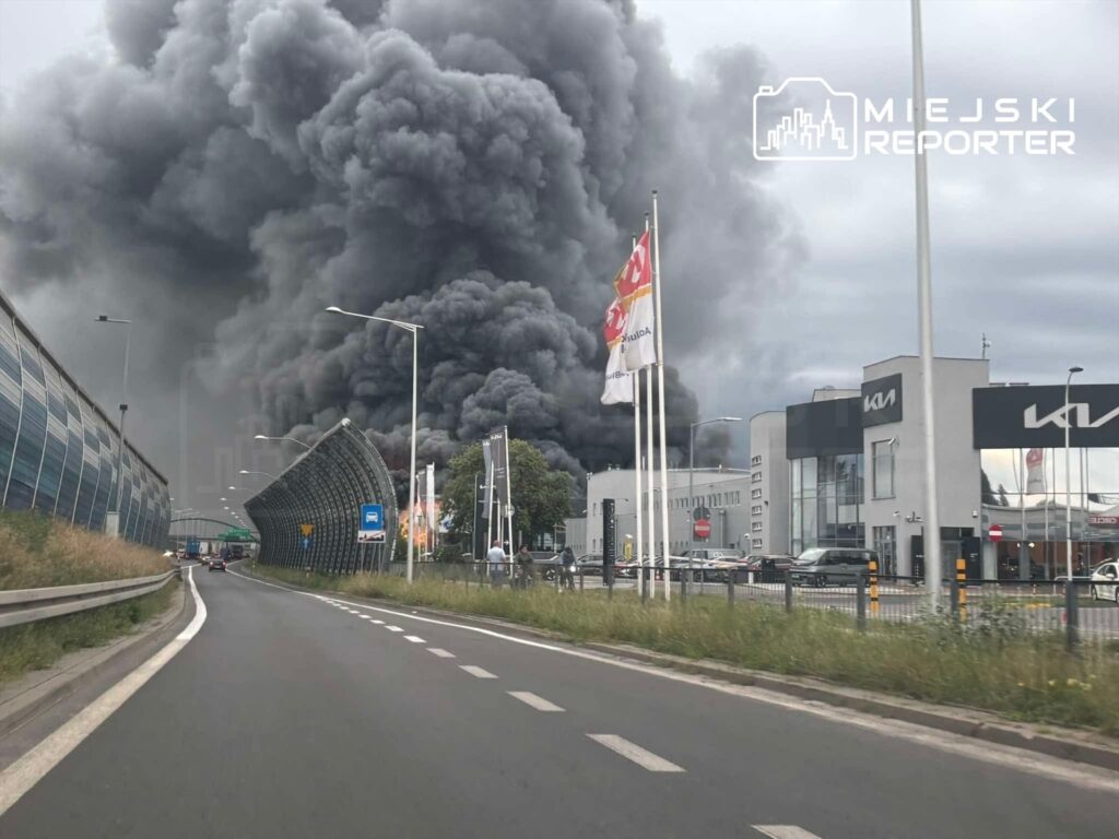 Kłęby czarnego dymu unoszą się nad nowoczesnym biurowcem i salonem samochodowym, widoczne z jezdni.