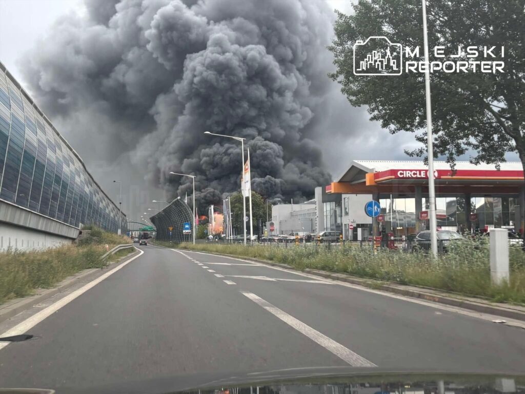 Gęste kłęby czarnego dymu unoszą się nad miastem, w tle widać salon samochodowy i stację benzynową.