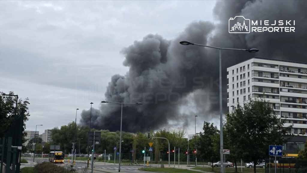 Gęste kłęby czarnego dymu unoszą się nad miastem, w tle nowoczesne budynki i przystanek autobusowy.