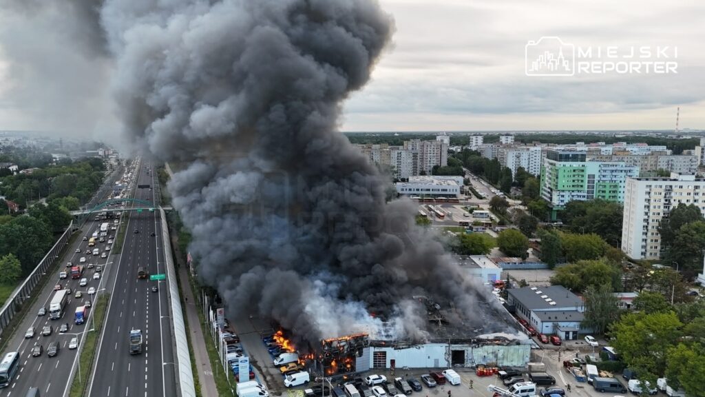 Wielkie kłęby czarnego dymu unoszą się nad płonącym salonem samochodowym, obok ruchliwej drogi.