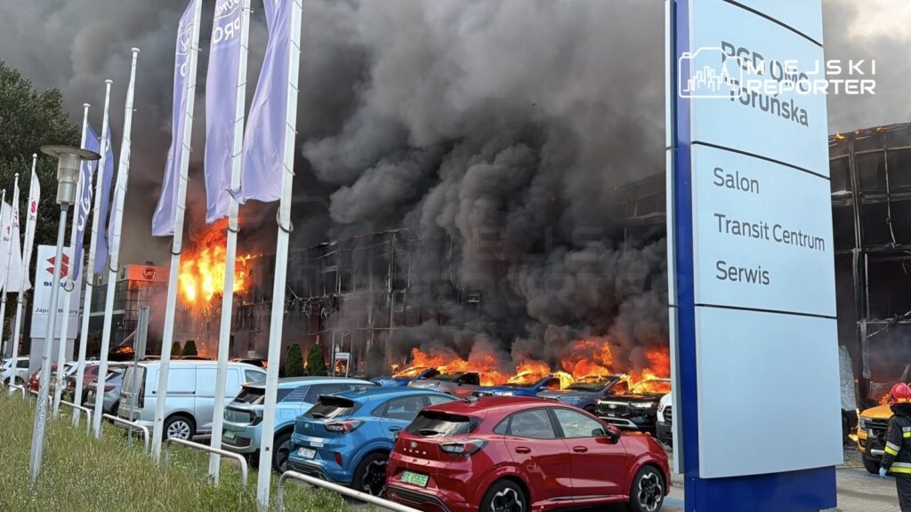 Ogromne płomienie i kłęby dymu wydobywają się z salonu samochodowego, w pobliżu zaparkowane pojazdy.