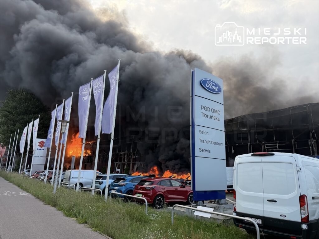 Czarne kłęby dymu unoszą się z płonącej hali, obok której stoją samochody i flagi marek motoryzacyjnych.