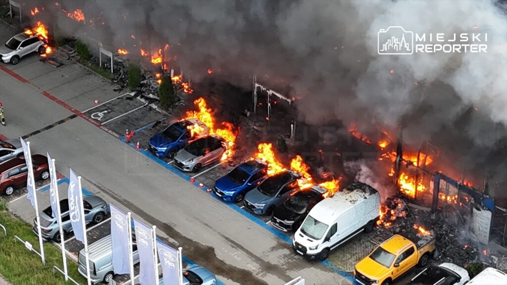Pojazdy zaparkowane na parkingu, otoczone płomieniami i dymem, podczas pożaru hali.