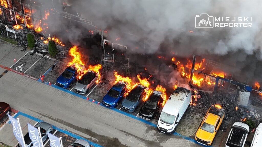 Płonąca hala z intensywnym ogniem i dymem, otaczająca zaparkowane samochody na parkingu.
