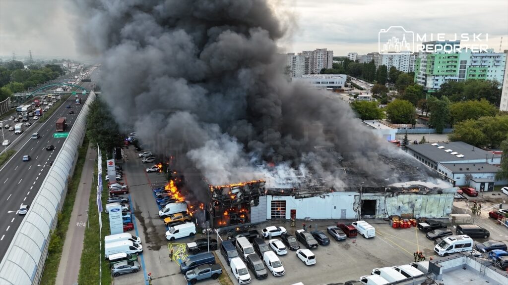 Hala przemysłowa objęta ogniem, z dymem unoszącym się w powietrze, w otoczeniu zaparkowanych pojazdów i ruchliwej drogi.