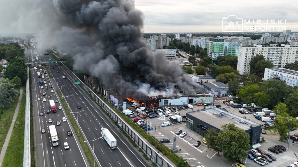 Pożar hali emitujący gęsty, czarny dym, z płomieniami widocznymi w ruinach budynku, obok ruchliwa autostrada.