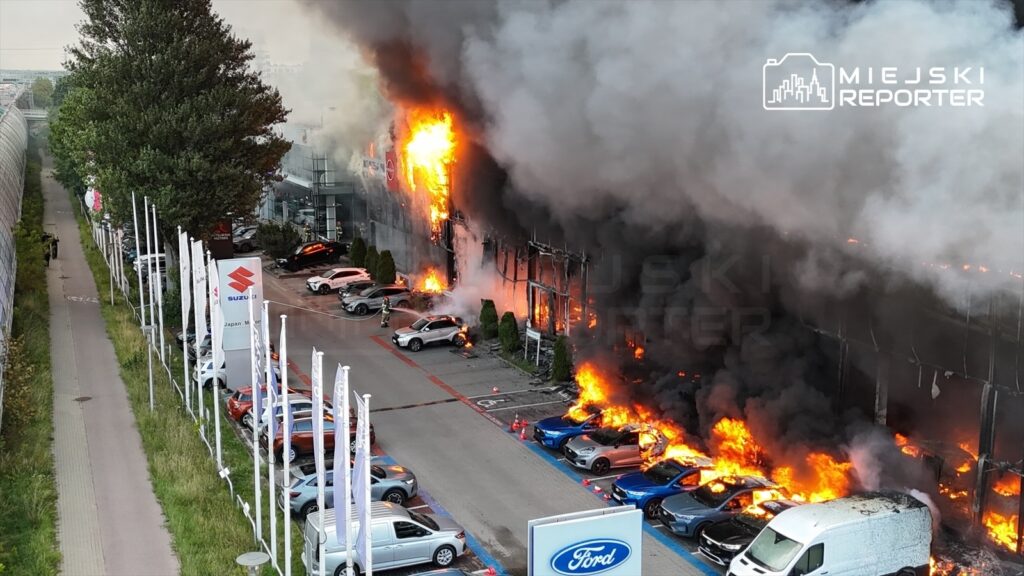 Hala w ogniu, z płomieniami i gęstym dymem unoszącym się nad zaparkowanymi samochodami.