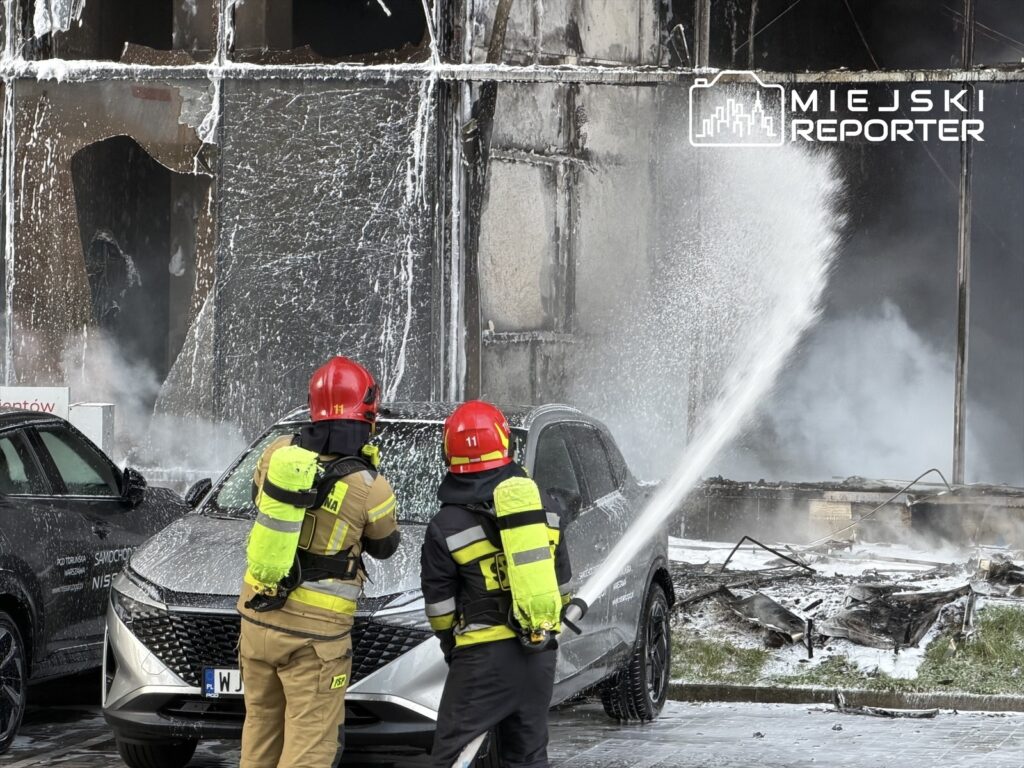 Strażacy w hełmach gaszą pożar hali, używając węża strażackiego, w otoczeniu zniszczonego budynku.
