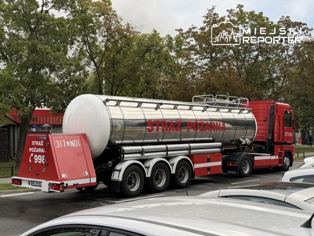 Czerwony wóz strażacki z metalowym zbiornikiem, oznaczony napisem "STRAŻ POŻARNA", parkuje na ulicy wśród innych samochodów.